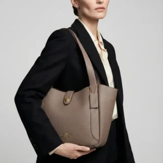 La Coach Hadley Turnlock Tote Bag en color Dark Stone es una cartera tote sofisticada y funcional, diseñada para acompañarte en los días más activos sin perder elegancia. Confeccionada en cuero de becerro Double Face de alta calidad, ofrece una estructura liviana, resistente y de acabado impecable. Su diseño minimalista incorpora el icónico cierre turnlock de Coach, que mantiene tus pertenencias seguras y le aporta un sello clásico y atemporal. El interior amplio permite llevar documentos, agenda, tablet o notebook pequeño, junto a tus esenciales diarios, mientras que el bolsillo interior con cierre facilita una mejor organización. Gracias a sus dos asas largas para el hombro, esta tote es cómoda para el uso diario y se adapta perfectamente tanto a looks de oficina como a outfits más relajados de fin de semana. El tono Dark Stone, combinado con herrajes dorados, la convierte en una pieza versátil y fácil de combinar durante todo el año. Disponible en Trendstore, 100% original, con envío a todo Chile y atención cercana. Una cartera Coach ideal si buscas calidad, diseño clásico y funcionalidad en un solo bolso.